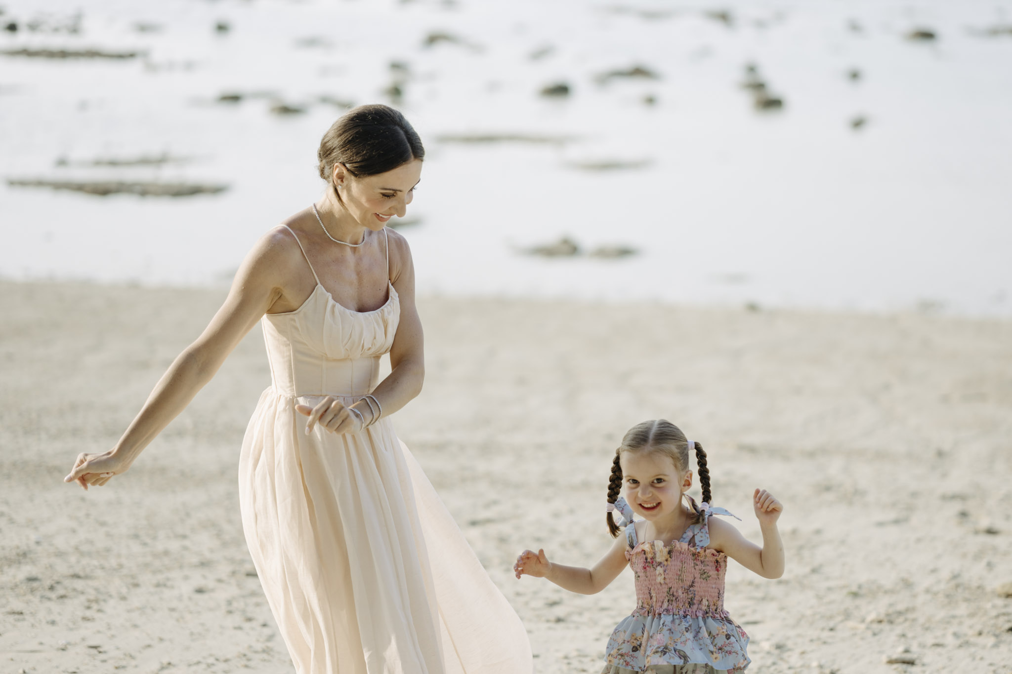 Soft, sunlit beach family photography session featuring parents and young daughter walking hand in hand on the sand, cuddling, laughing, and sharing tender candid moments by the ocean and tropical greenery in a natural, airy lifestyle style.
