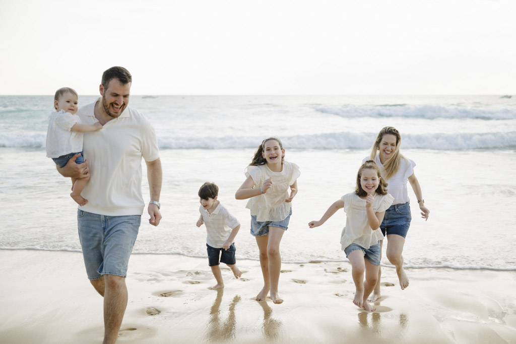 Light-filled coastal family photography session capturing joyful, candid moments of parents and children running barefoot on the beach in coordinated white and denim outfits with soft ocean waves in the background.